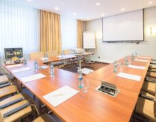 Bright conference room with U-shaped wooden tables, leather chairs, water bottles, projector, and natural light from large windows