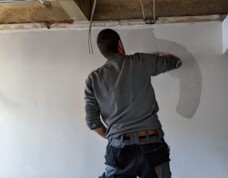 Worker smoothing plaster on a hotel room wall during renovation, with exposed ceiling and wiring visible