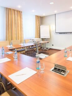 Heller Konferenzraum mit U-förmigen Holztischen, beigen Lederstühlen, Wasserflaschen, Notizblöcken, Beamer und viel Tageslicht