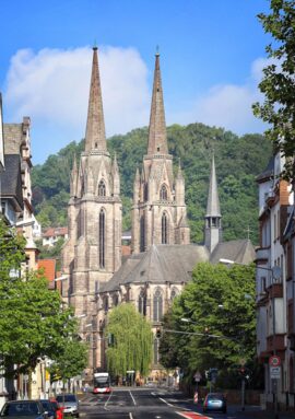 Historic Gothic church with twin tall spires on a tree-lined city street, backed by lush green hillside under blue sky