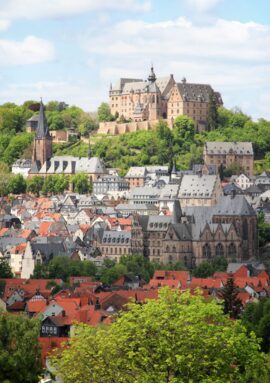 Historic castle atop lush green hill overlooking red-roofed town with medieval churches under blue sky