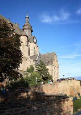 Historic stone castle with clock tower under clear sky, surrounded by greenery and guests relaxing on stone terraces