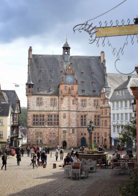 Historic town square with stone building featuring a clock tower, outdoor café seating, and visitors enjoying the lively atmosphere