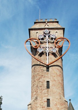 Historic stone tower with decorative neon heart and floral design against a clear blue sky, surrounded by leafy trees