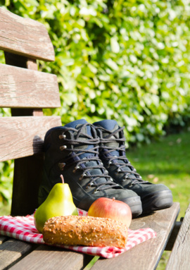 Outdoor hiking boots on a wooden bench next to a pear, apple, and multigrain bread on a red checkered cloth