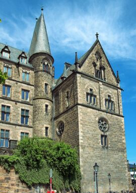 Historic stone building with Gothic architectural details including a tall conical tower and pointed arch windows under a clear blue sky