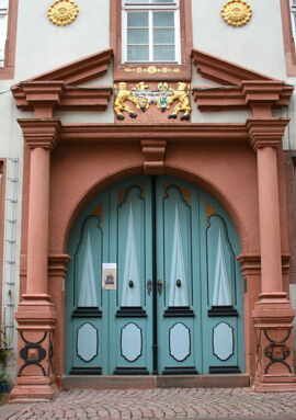 Historic building entrance with ornate blue double doors framed by red stone columns and golden decorative accents, flanked by benches.