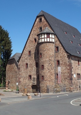Historic stone building with steep slate roof and small windows housing a museum, set beside a quiet street under clear blue sky