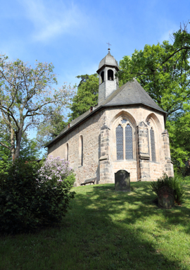Historic stone chapel with Gothic arched windows nestled among green trees and flowering bushes on a sunny day