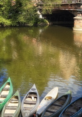 Row of colorful canoes moored on calm river water beneath a stone bridge surrounded by green trees