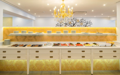 Elegantes Frühstücksbuffet mit verschiedenen Käsesorten, Aufschnitt, Früchten und Tellern unter einem goldenen Kronleuchter in hellem Speiseraum