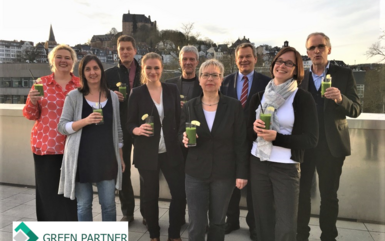 Neun Personen genießen grüne Smoothies auf einer Hoteldachterrasse mit Blick auf historische Stadt und Schloss bei Sonnenuntergang