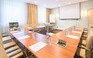 Heller Konferenzraum mit U-förmigen Holztischen, beigen Lederstühlen, Wasserflaschen, Notizblöcken, Beamer und viel Tageslicht