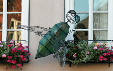 Große dekorative Metallinsekten-Skulptur an Hotelzimmerfenster mit lebhaften rosa und roten Blumen im Blumenkasten