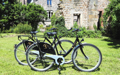 Zwei schwarze Gäste-Fahrräder auf grüner Wiese neben einem historischen Steinbau mit Kletterpflanzen
