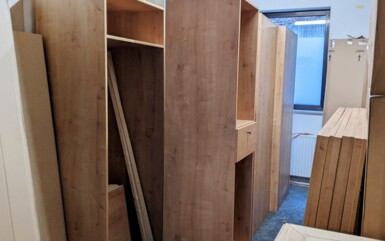 Unassembled wooden furniture panels and cabinets stacked in a storage room with a small window and carpeted floor