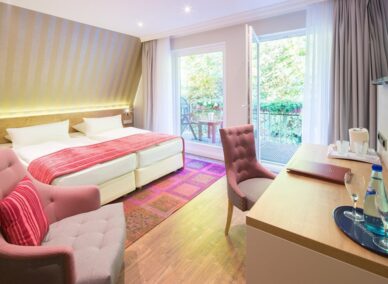 Bright hotel room with twin beds, pink accents, seating area, balcony with garden view, desk with water and glasses