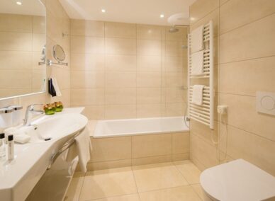 Modernes Hotelbad mit beigen Fliesen, geräumiger Badewanne, weißem Waschbecken, beheiztem Handtuchhalter und sorgfältig angeordneten Pflegeartikeln