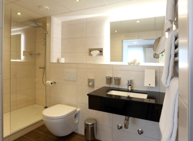 Modernes Hotelbad mit begehbarer Dusche, wandhängendem WC, schwarzer Waschtischplatte, großem beleuchtetem Spiegel und frischen Handtüchern