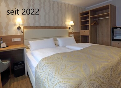 Gemütliches Hotelzimmer mit zwei Einzelbetten und gemustertem Bettüberwurf, Holzmöbeln, Schreibtisch, Minibar und warmem Nachtlicht