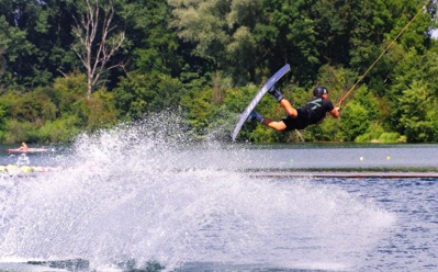 Gast beim Wakeboarden auf einem See, umgeben von dichtem Grün, genießt aktiven Wassersport an sonnigem Tag