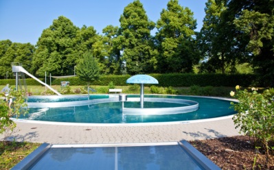 Runder Außenpool mit Wasserrutsche und Pilzbrunnen, umgeben von Bäumen und gepflasterter Terrasse bei Sonnenschein