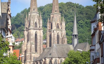 Historische gotische Kirche mit zwei hohen Türmen an einer von Bäumen gesäumten Straße, im Hintergrund grüner Hügel unter blauem Himmel