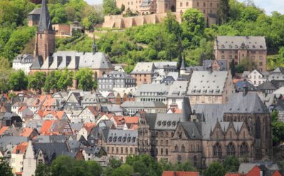 Historisches Schloss auf grünem Hügel mit Blick auf rotgedeckte Stadt und mittelalterliche Kirchen bei blauem Himmel