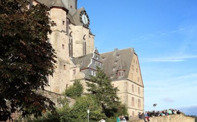 Historische Steinschloss mit Uhrenturm unter klarem Himmel, umgeben von Grün und Gästen auf Steinterrassen