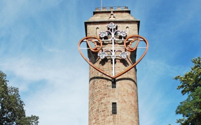 Historischer Steinturm mit dekorativem Neonherz und Blumendesign vor klarem blauem Himmel, umgeben von Laubbäumen