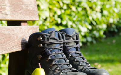 Wanderschuhe auf einer Holzbank neben einer Birne, einem Apfel und mehrkörnigem Brot auf rotem Karotuch im Freien