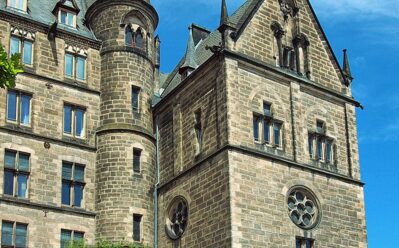 Historisches Steingebäude mit gotischen Details, spitzem Turm und Bogenfenstern unter klarem blauem Himmel