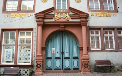 Historischer Hauseingang mit verzierten blauen Doppeltüren, roten Steinsäulen und goldenen Dekorationen, seitlich Sitzbänke.