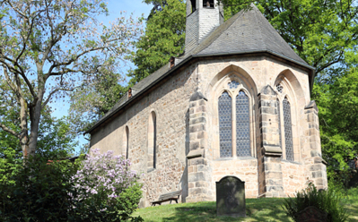 Historische Steinkapelle mit gotischen Bogenfenstern, umgeben von grünen Bäumen und blühenden Büschen an einem sonnigen Tag