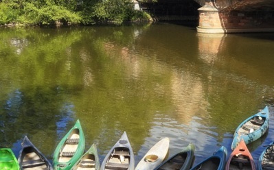 Reihe bunter Kanus auf ruhigem Flusswasser unter einer Steinbrücke, umgeben von grünen Bäumen
