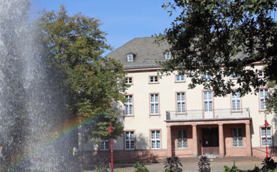 Historisches Hotelgebäude mit klassischer Architektur, umgeben von üppigen Gärten und einem großen Springbrunnen mit Regenbogen im Wassersprühnebel