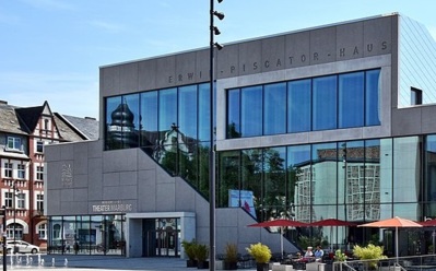 Modernes Theatergebäude mit großen Glasfenstern, Außenbestuhlung mit roten Sonnenschirmen und Spiegelungen historischer Architektur