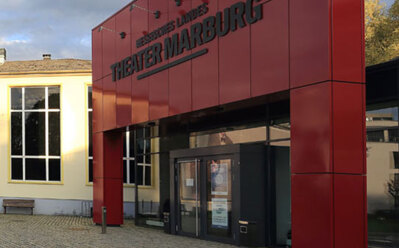 Moderner Theater-Eingang mit auffälliger roter Verkleidung und großen Glastüren auf Kopfsteinpflaster unter bewölktem Himmel