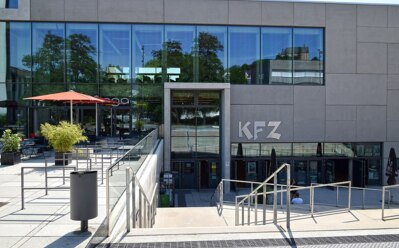 Moderne Hotelfassade mit großen Glasfenstern, Außensitzbereichen unter roten Sonnenschirmen und barrierefreien Zugängen zum Eingang