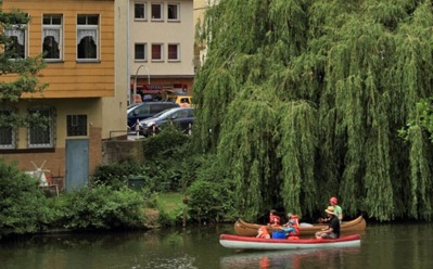 Gäste beim Kanufahren und Stand-up-Paddeln auf ruhigem Fluss, umgeben von üppigem Grün und Gebäuden