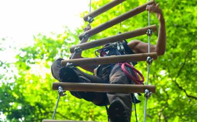 Gast klettert auf einer Seilleiter im Outdoor-Abenteuerparcours umgeben von dichtem grünen Blätterdach