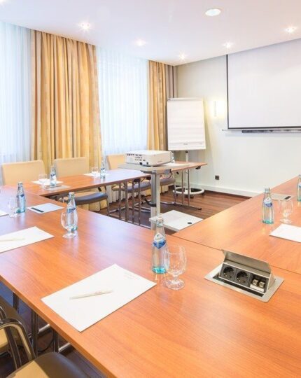 Heller Konferenzraum mit U-förmigen Holztischen, Ledersesseln, Wasserflaschen, Beamer und viel Tageslicht durch große Fenster