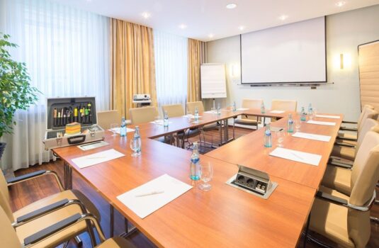 Bright conference room with U-shaped wooden tables, beige chairs, bottled water, writing pads, projector, and flip chart for meetings.