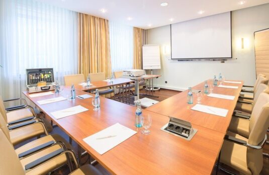 Bright conference room with U-shaped wooden tables, leather chairs, water bottles, projector, and natural light from large windows