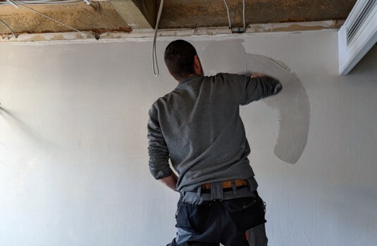 Worker smoothing plaster on a hotel room wall during renovation, with exposed ceiling and wiring visible