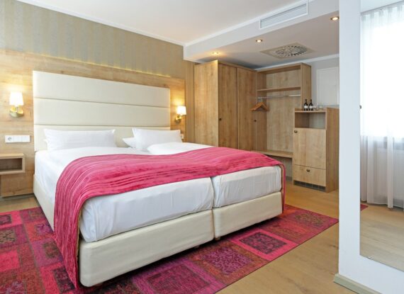 Modernes Hotelzimmer mit zwei zusammengestellten Betten, pinker Tagesdecke, Holzkleiderschrank, weicher Beleuchtung und großem Fenster mit Gardinen