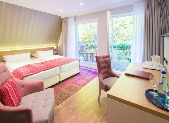 Helles Hotelzimmer mit zwei Einzelbetten, rosa Akzenten, Balkon mit Blick ins Grüne und Schreibtisch mit Wasserflasche und Gläsern