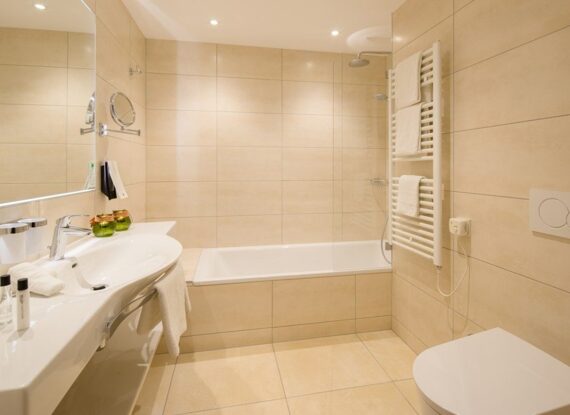 Modernes Hotelbad mit beigen Fliesen, geräumiger Badewanne, weißem Waschbecken, beheiztem Handtuchhalter und sorgfältig angeordneten Pflegeartikeln