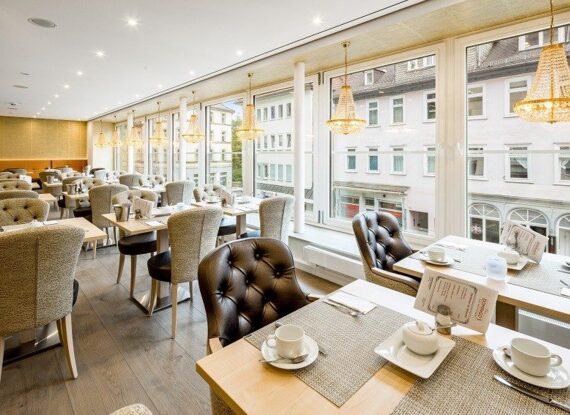 Helles Hotelrestaurant mit gepolsterten Stühlen, großen Fenstern mit Blick auf Stadtgebäude und eleganten Kronleuchtern an der Decke
