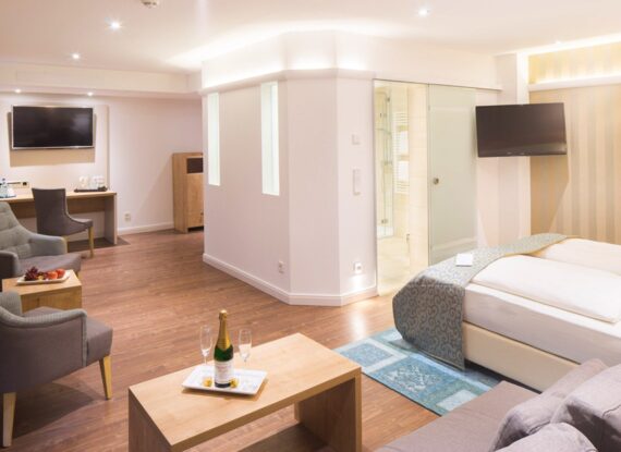 Geräumige, moderne Hotel-Suite mit Doppelbett, Sitzbereich, Flachbildfernsehern, Holzboden und Bad mit gläserner Duschabtrennung.